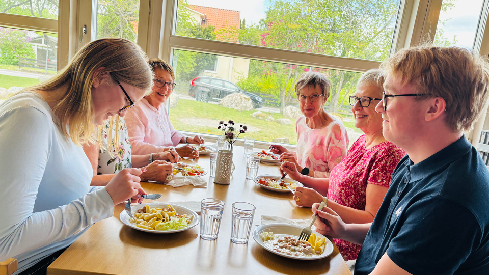 Elever och seniorer sitter vid bord och äter lunch i skolmatsal. Foto.