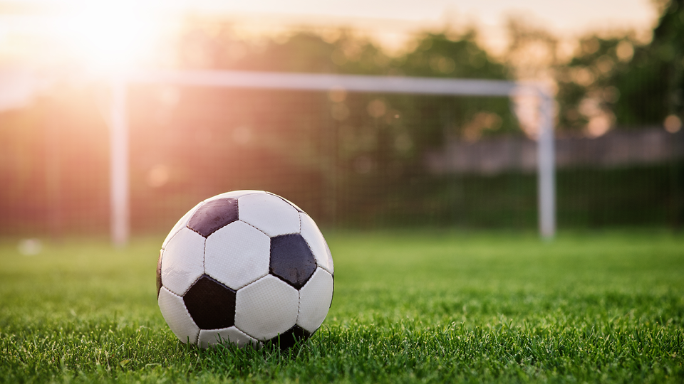 En tjej står med fotboll och ler in i kameran. I bakgrunden kickar två andra tjejer boll.